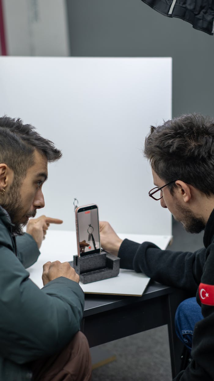 Two young men discussing photography techniques while examining a phone setup indoors.